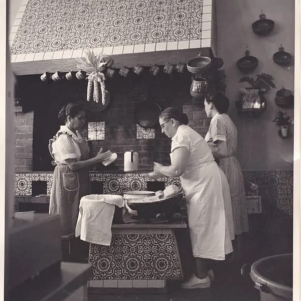 Three women preparing food in a traditional Mexican kitchen with decorative tiles and hanging pots.
