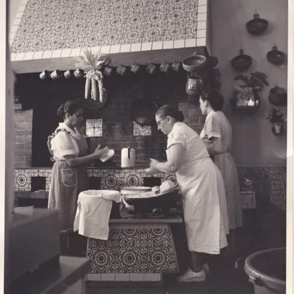 Three women preparing food in a traditional Mexican kitchen with decorative tiles and hanging pots.