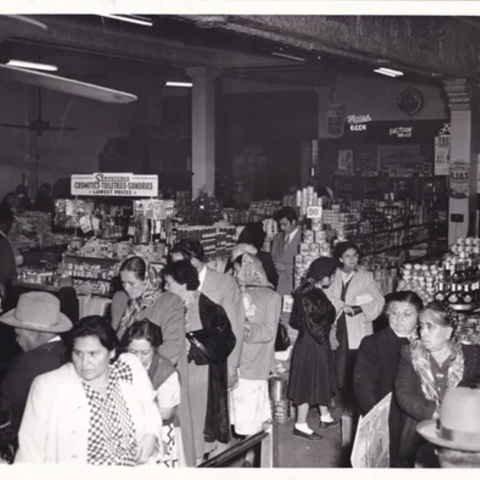 Black and white photo of a busy grocery store with many shoppers and shelves stocked with products.