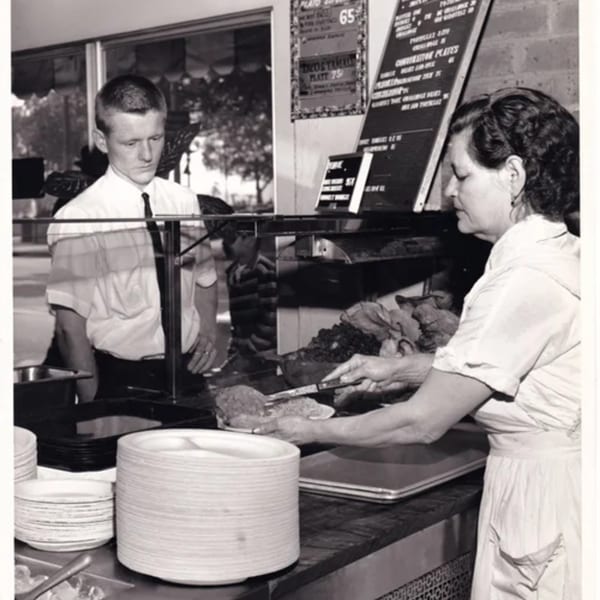 Black and white photo of a woman serving food to a man at a La Luz del Día counter.