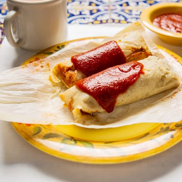 Two La Luz del Día tamales topped with red sauce on a yellow plate with a cup and salsa.