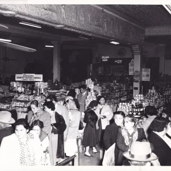 Black and white photo of a busy grocery store with many customers browsing shelves of products.