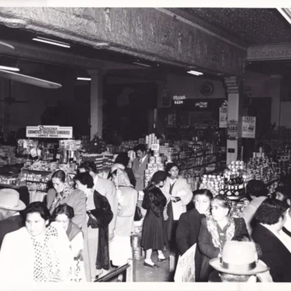 Black and white photo of a busy grocery store with many customers browsing shelves of products.