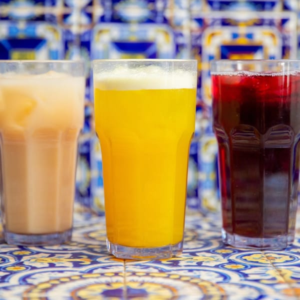 Three colorful Mexican aguas frescas: horchata, orange, and jamaica, on a blue and yellow tiled surface.
